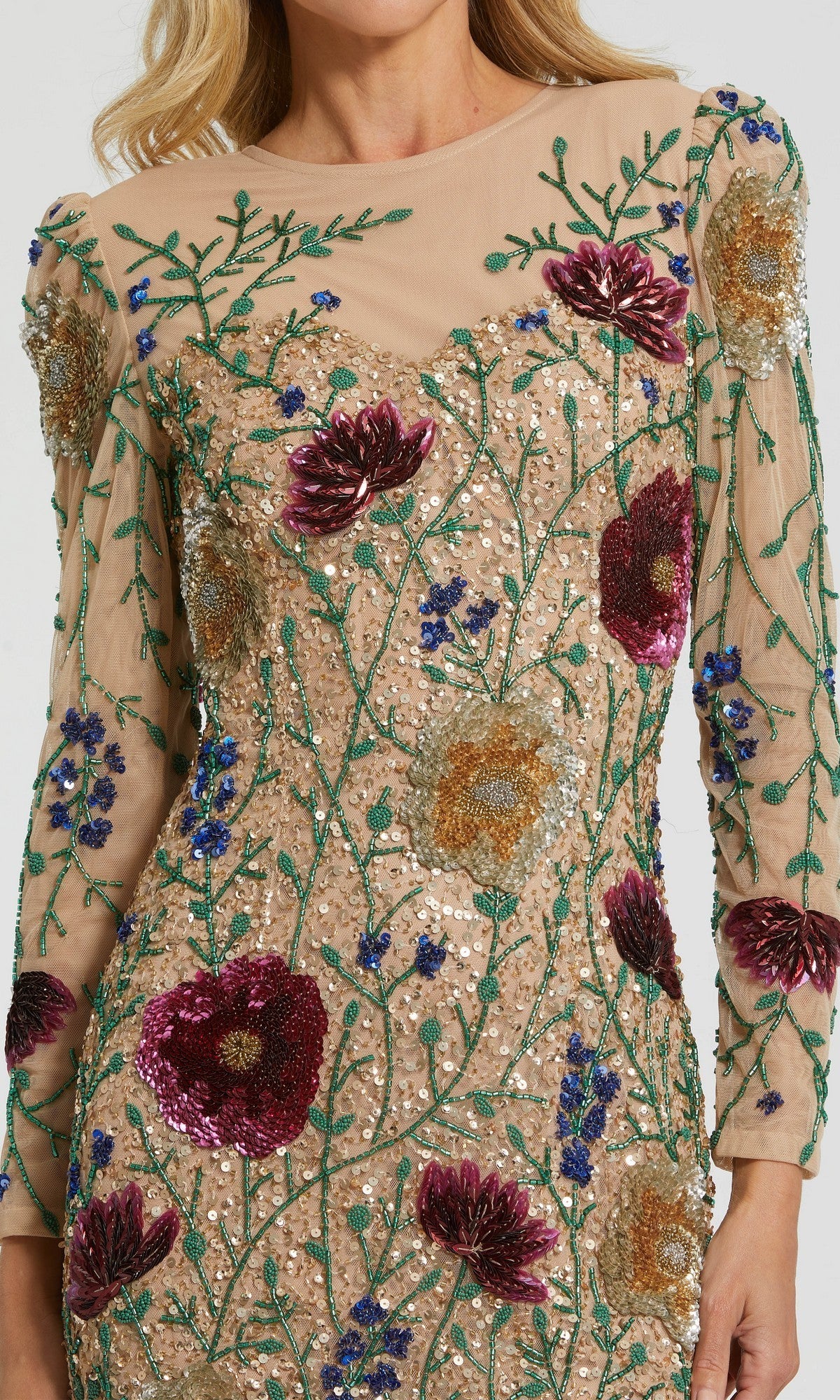 A woman wears the Short Party Dress: Mac Duggal 24006, a sheer, long-sleeve mini dress featuring sequin floral embroidery in red, gold, green, and blue on a nude background. The beaded floral design is visible on her upper torso and arms.